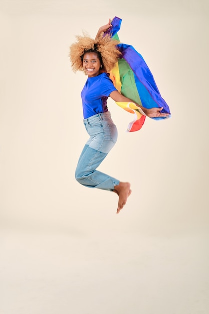 Mulher afro feliz e animada, saltando com uma bandeira de orgulho lgbt de arco-íris em um fundo isolado. Direitos iguais e conceito de apoio à comunidade LGBT.