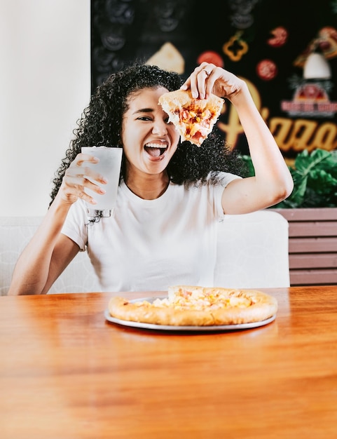 Foto mulher afro feliz mostrando uma fatia de pizza em um restaurante mulher afro sorridente saboreando uma pizza em um restaurante