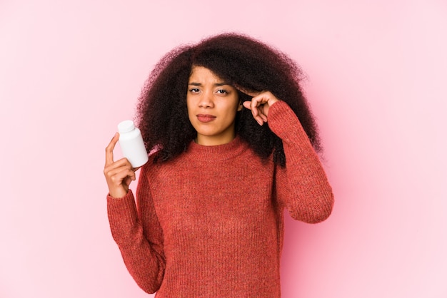 Mulher afro nova que mantém vitaminas isoladas Mulher afro nova que prende vitaminasshowing um gesto do desapontamento com dedo indicador.