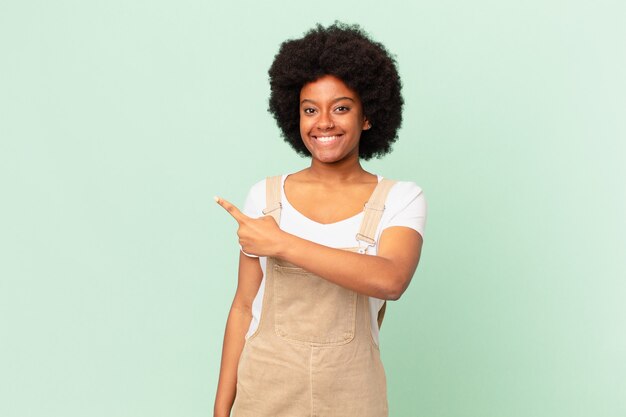 Mulher afro sorrindo alegremente, sentindo-se feliz e apontando para o lado e para cima, mostrando o objeto no conceito do chef do espaço de cópia