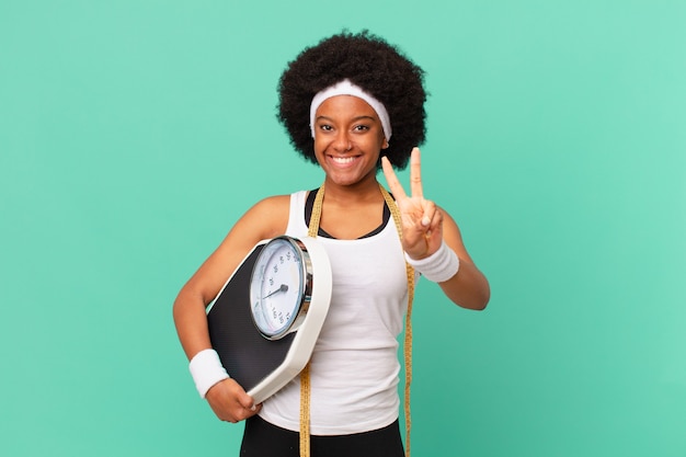 Mulher afro sorrindo e parecendo feliz, despreocupada e positiva, gesticulando vitória ou paz com o conceito de dieta de uma mão