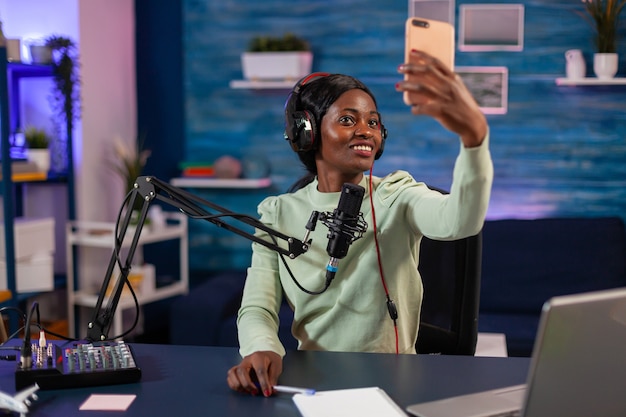 Mulher afro tirando selfie com smartphone e usando equipamento profissional para registrar o episódio na sala de estar. Produção on-line no ar, apresentador de podcast de show de internet transmitindo conteúdo ao vivo, gravando soc digital