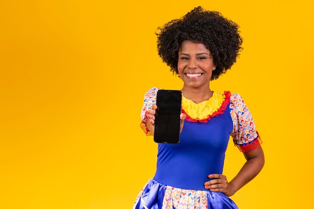 Mulher afro vestida com roupa de festa junina com um smartphone com a tela branca para o texto