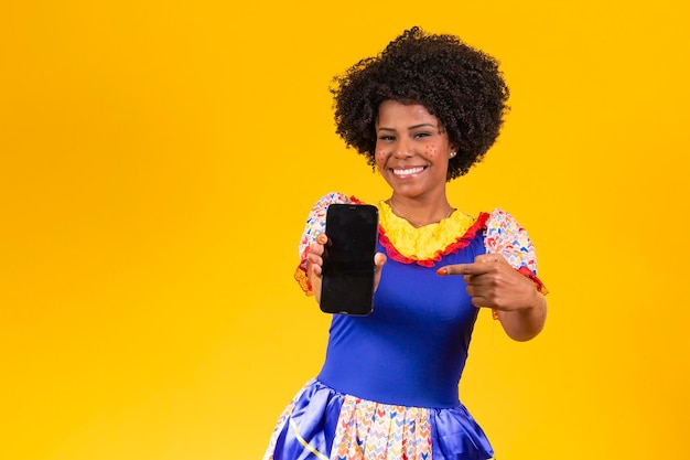 Mulher afro vestida com roupa de festa junina com um smartphone com a tela branca para o texto