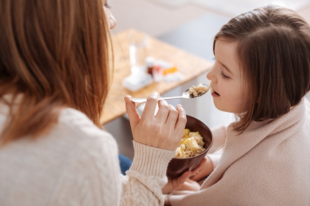 Mulher agradável e adorável segurando uma colher e dando café da manhã para sua filhinha