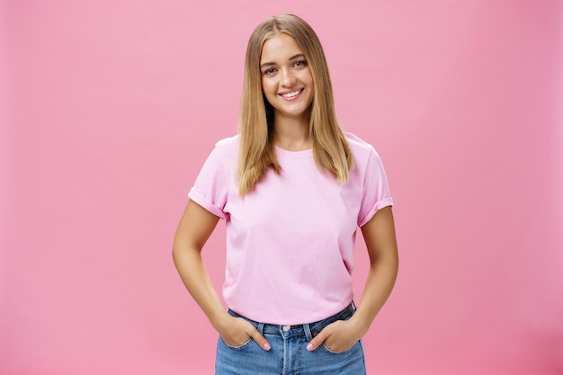 Mulher agradável e charmosa com rosto rechonchudo e cabelo loiro em uma camiseta casual sorrindo amplamente de mãos dadas nos bolsos sendo simpática e simpática olhando despreocupada e calma