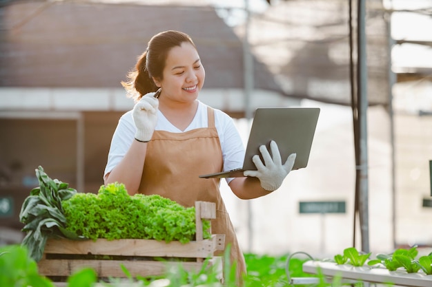 Mulher agricultora asiática trabalhando com laptop em fazenda hidropônica de vegetais orgânicos. proprietário do jardim de salada hidropônica verificando a qualidade do vegetal na plantação de estufa.