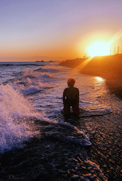 Mulher ajoelhada na praia contra o céu durante o pôr do sol
