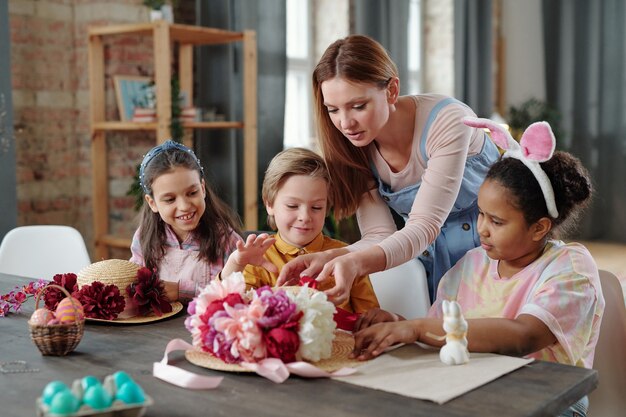Foto mulher ajudando crianças a fazer artesanato enquanto elas estão sentadas à mesa decorando chapéus com flores