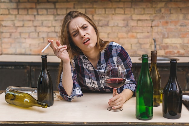 Foto mulher alcoólica triste e desperdiçada nova que senta-se no sofá da cozinha que bebe o vinho vermelho que guarda o vidro completamente bêbado que olha deprimido só e sofrendo da ressaca no alcoolismo e no abuso de álcool.