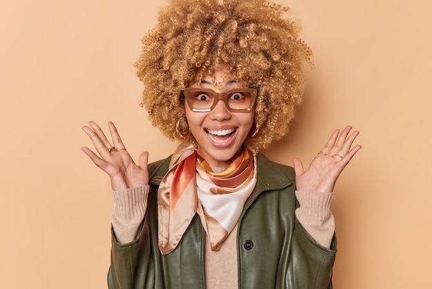 Mulher alegre animada com cabelo loiro encaracolado mantém as palmas das mãos levantadas exclama de alegria reage a notícias agradáveis vestidas com roupa da moda isolada sobre fundo marrom Conceito de reações humanas