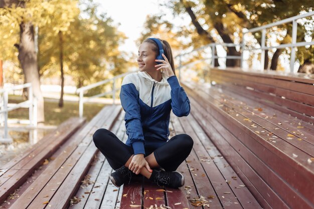 Mulher alegre apta no sportswear sentado em suportes de madeira e ouvindo música em fones de ouvido.