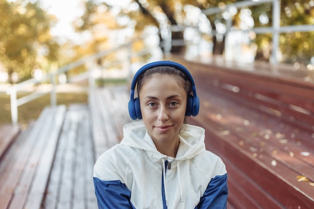 Mulher alegre apta no sportswear sentado em suportes de madeira e ouvindo música em fones de ouvido.
