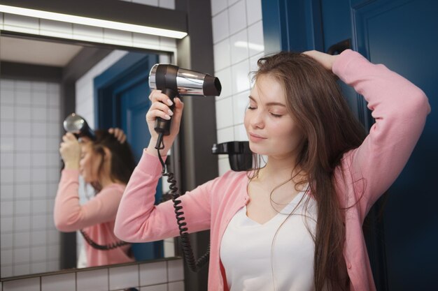 Mulher alegre atraente sorrindo enquanto seca o cabelo com um secador de cabelo