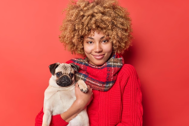 Mulher alegre bonita com cabelo loiro encaracolado usa jumper casual e cachecol no pescoço posa com cachorro vai passear com animal de estimação favorito isolado sobre fundo vermelho. Conceito de animais de pessoas