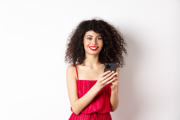 Mulher alegre com cabelo encaracolado, usando o smartphone com um vestido vermelho, sorrindo para a câmera, em pé sobre um fundo branco. copie o espaço