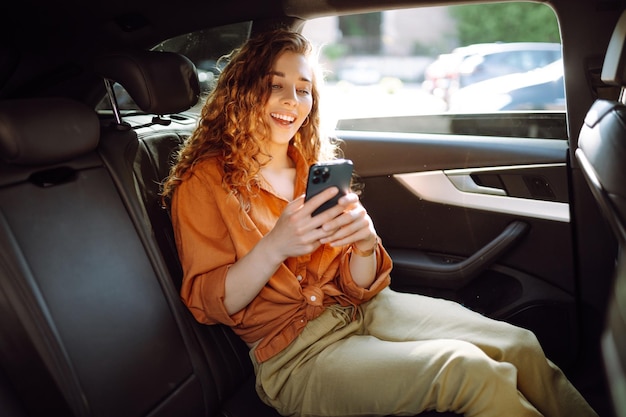 Mulher alegre com cabelos encaracolados e roupas elegantes senta-se no banco de trás de um carro usa um telefone celular