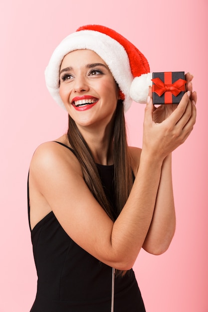 Mulher alegre com chapéu de Natal segurando uma caixa de presente isolada sobre fundo rosa