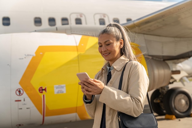 Mulher alegre com telefone em pé ao ar livre no aeroporto perto de avião