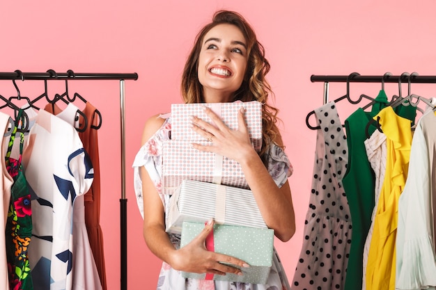 mulher alegre com um vestido em pé na loja perto do cabideiro com caixas de presentes isoladas em rosa