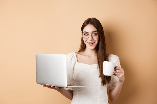 Mulher alegre de óculos, bebendo café e trabalhando no laptop, sorrindo para a câmera, de pé sobre um fundo bege.