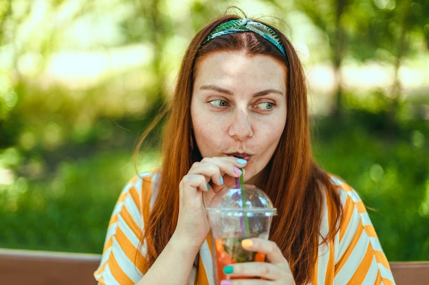 Mulher alegre de verão com coquetel gelado sentada em um banco de madeira no parque da cidade