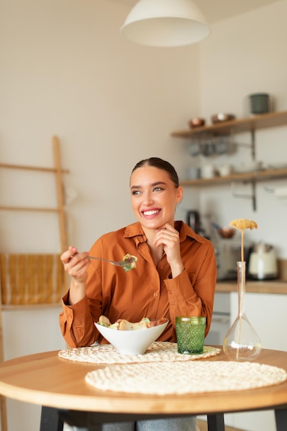 Foto mulher alegre, desfrutando de uma deliciosa salada césar, sentada à mesa na cozinha, olhando de lado e sorrindo