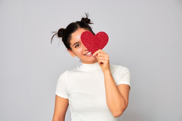 Foto mulher alegre e apaixonada por um penteado engraçado segurando um pequeno coração vermelho cintilante do dia dos namorados e cobrindo um olho enquanto sorri