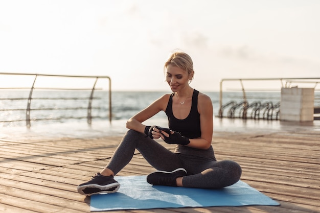 Mulher alegre e fitness sentada no tapete e usando um smartphone