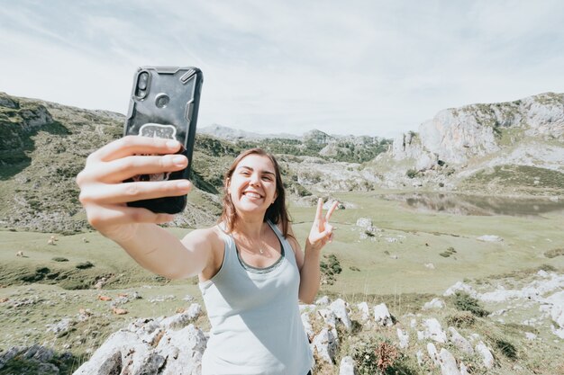 Mulher alegre e simpática sorrindo e fazendo selfie enquanto descansa nas colinas da montanha, dia claro nos lagos no topo das montanhas, símbolo da paz