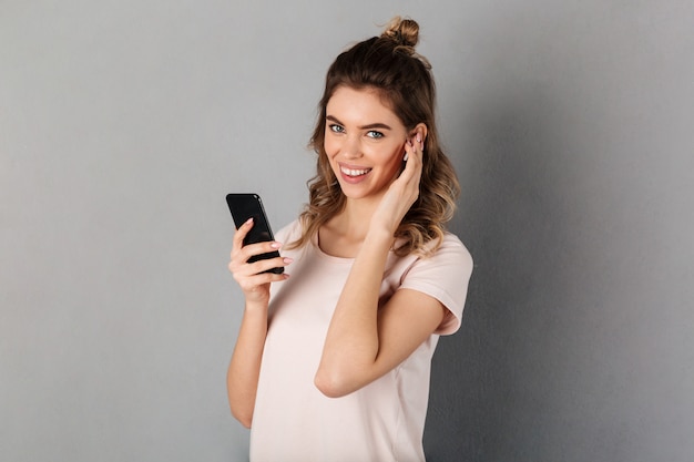 Mulher alegre em ouvir música de t-shirt do smartphone com fones de ouvido enquanto mais cinza