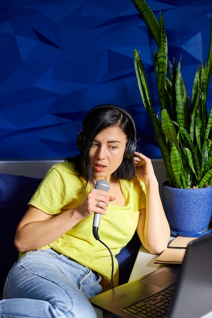 Mulher alegre em roupas casuais Gravando um podcast, falando em um microfone com fones de ouvido e laptop, notebook