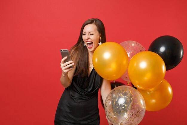 Mulher alegre em um vestido preto gritando segurando balões de ar, usando telefone celular, enquanto comemora isolado sobre fundo vermelho. Dia dos namorados feliz ano novo aniversário maquete feriado festa conceito.