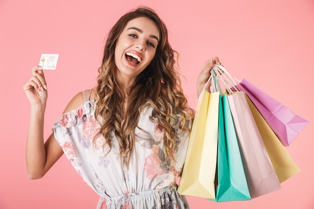 Mulher alegre em um vestido segurando um cartão de crédito e sacolas de compras coloridas, isoladas em rosa