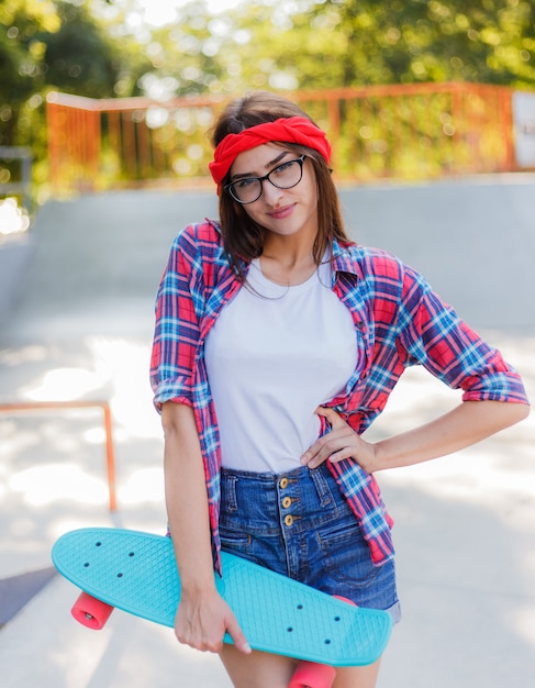 Mulher alegre emocional jovem hippie, vestida com roupas elegantes mantém o skate na mão no skatepark em dia ensolarado