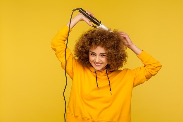 Mulher alegre extravagante usando ferro de ondulação para penteado afro perfeito garota na moda passando cachos de cabelo aliciamento com ferramenta profissional conceito de cuidados de beleza tiro de estúdio interno isolado em fundo amarelo