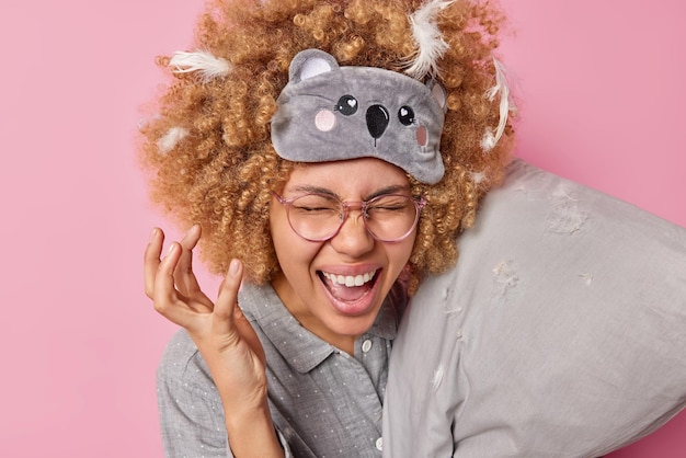 Mulher alegre mantém a mão levantada exclama alegremente estar de bom humor durante a manhã tem cabelo encaracolado com penas presas segura travesseiro vestido com roupa de dormir isolada sobre fundo rosa Hora de descansar