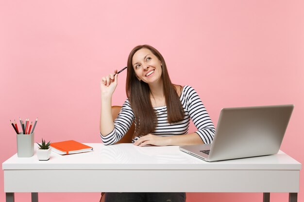 Mulher alegre segurando um lápis perto da cabeça, olhando para cima, pense sonhando, sente-se, trabalhe em uma mesa branca com um laptop pc contemporâneo