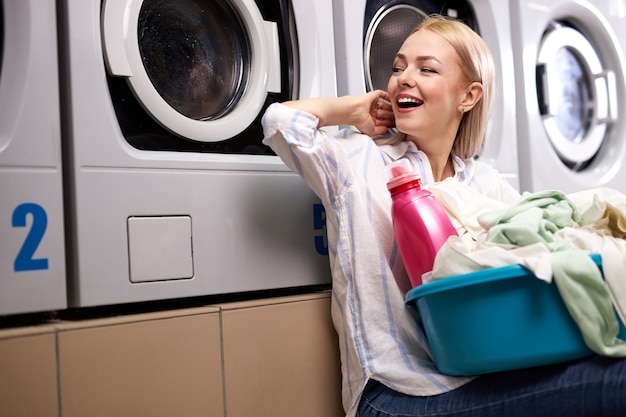 Mulher alegre sentada feliz esperando o fim da lavanderia, sorrindo, segurando a bacia com roupas sujas e um frasco de detergente rosa