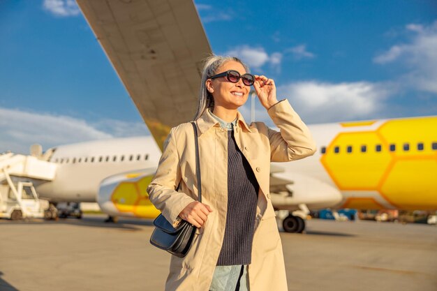Mulher alegre tocando óculos de sol e sorrindo em pé no aeródromo com avião de passageiros no fundo