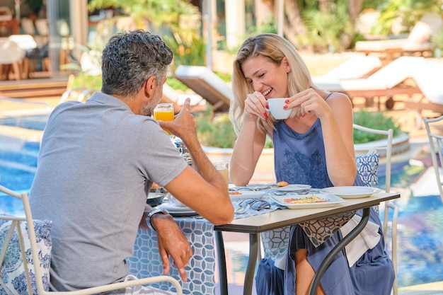Mulher alegre tomando uma xícara de café enquanto passa tempo com o homem bebendo suco na mesa no resort perto da piscina em dia ensolarado