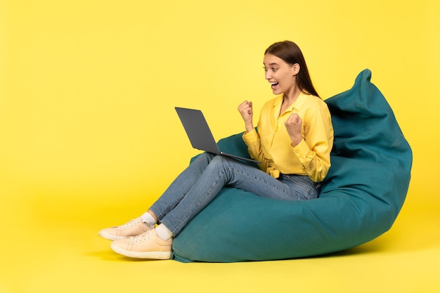 Mulher alegre usando laptop gesticulando sim sentado sobre fundo amarelo