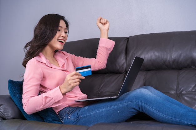 Mulher alegre usando laptop para fazer compras on-line com cartão de crédito na sala de estar bem sucedida