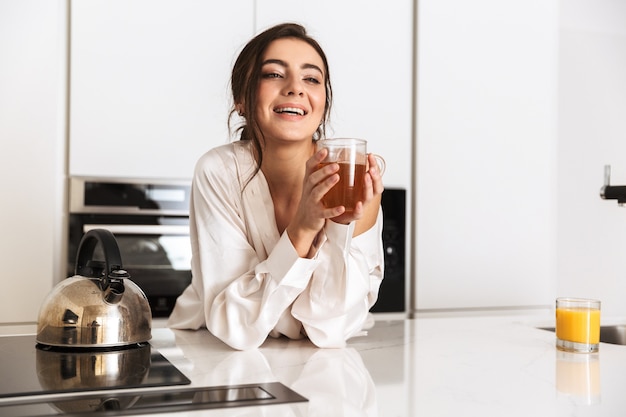 mulher alegre vestindo roupas de seda sorrindo, enquanto bebe chá na cozinha de casa