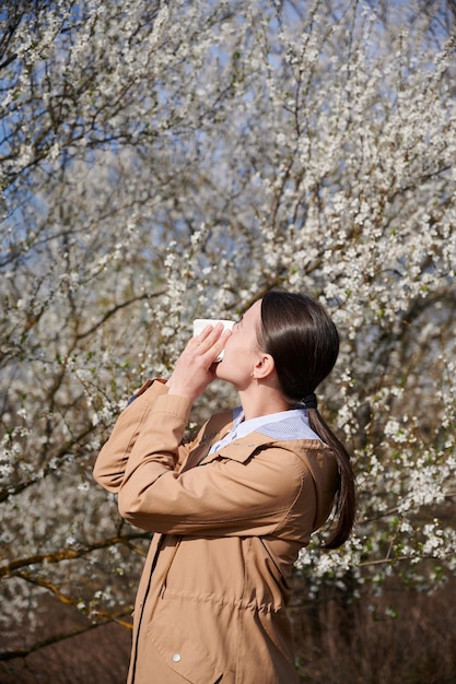 Mulher alérgica que sofre de alergia sazonal na primavera posando em um jardim em flor na primavera Mulher jovem espirrando e soprando o nariz na frente de uma árvore em flor conceito de alergia de primavera