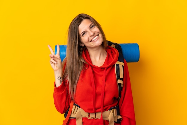 Mulher alpinista eslovaca com uma mochila grande isolada em um fundo amarelo sorrindo e mostrando sinal de vitória