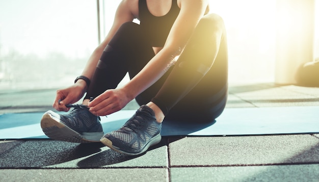 Mulher amarrando o cadarço de um tênis esportivo sentada em um tapete na academia