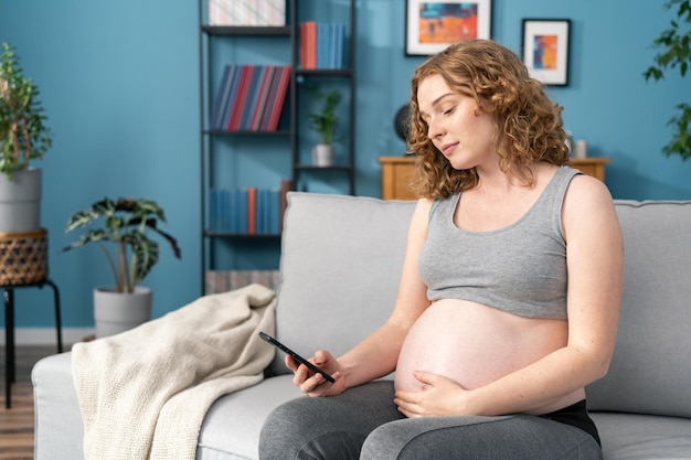 Mulher americana caucasiana grávida feliz com smartphone em casa