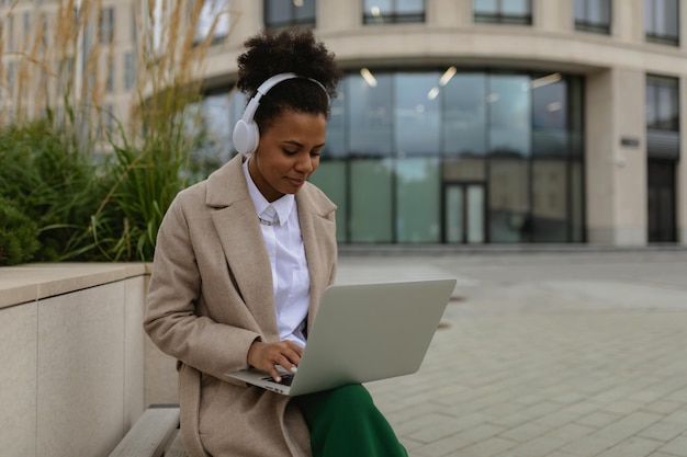 Mulher americana elegante trabalhando no laptop com fones de ouvido fora do escritório