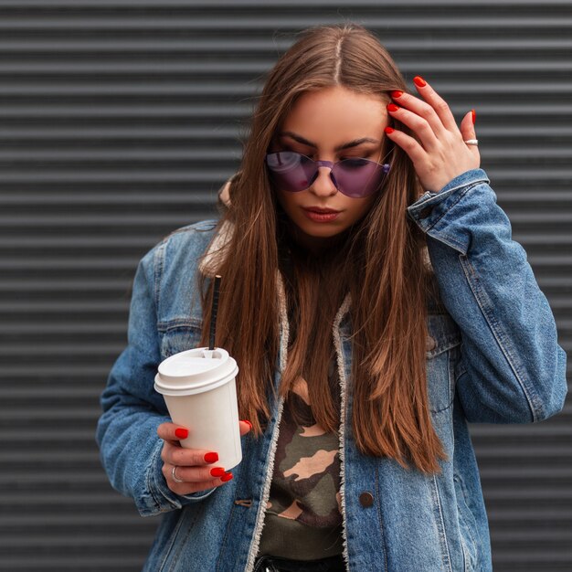Mulher americana jovem hippie em elegantes óculos roxos glamourosos em uma jaqueta jeans azul da moda com café alisa o cabelo e olhando para baixo perto da parede de metal vintage. Modelo de moda linda garota ao ar livre.